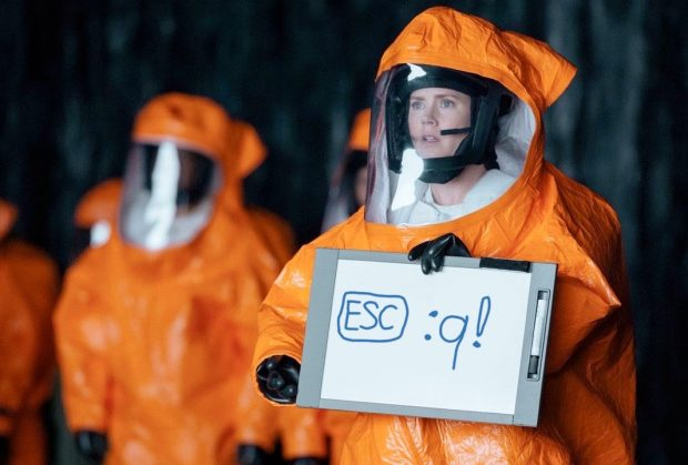 Louise Banks (Amy Adams) in Arrival (2016) wearing a protective suit holding a sign with handwriting saying: ESC :q!