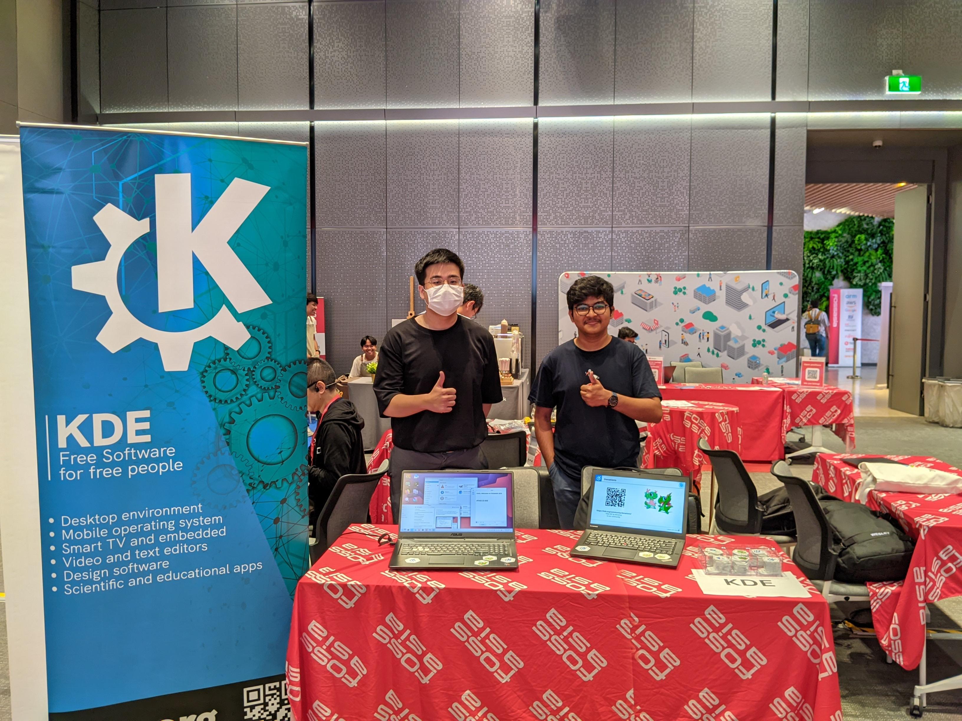 A view of KDE's booth at #FOSSASIA. On a table with a red tablecloth printed with the FOSSASIA logo are two laptops showing KDE software. Two young men stand behind the table doing the "thumbs up" sign. On the left is a bright blue banner with the KDE logo.